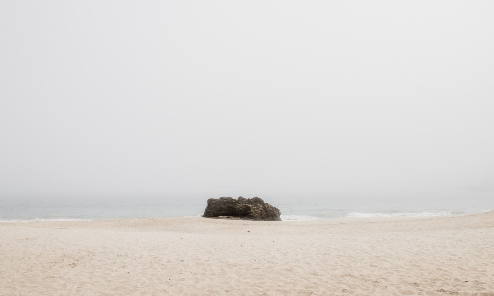 Roca oscura solitaria en la orilla, cielo blanco de niebla, arena clara y vacía. Agua #P2, fotografía de Cristian Letelier, edición limitada