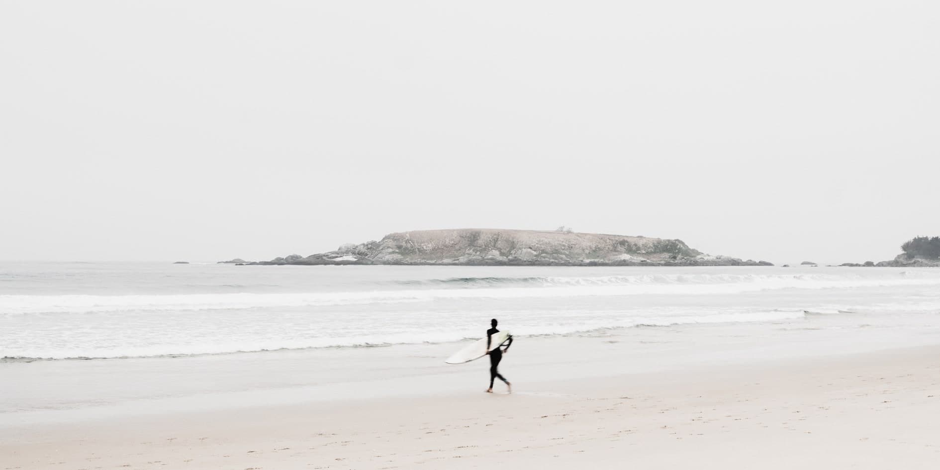 Surfista caminando con tabla por la orilla, isla rocosa al fondo, tonos pálidos. Agua #P4, fotografía de Cristian Letelier, edición limitada