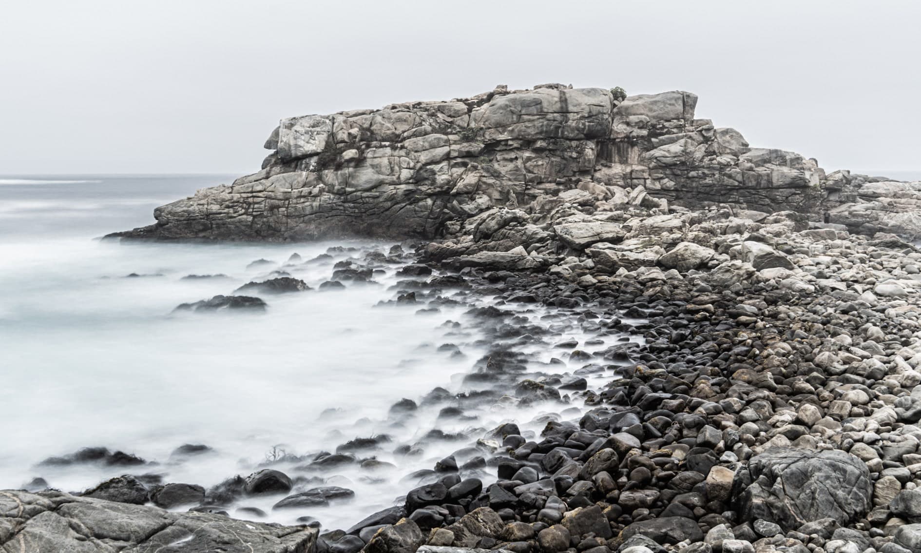 Roquerío gris con piedras redondas, mar sedoso en larga exposición, cielo blanco. Agua #P14, fotografía de Cristian Letelier, edición limitada
