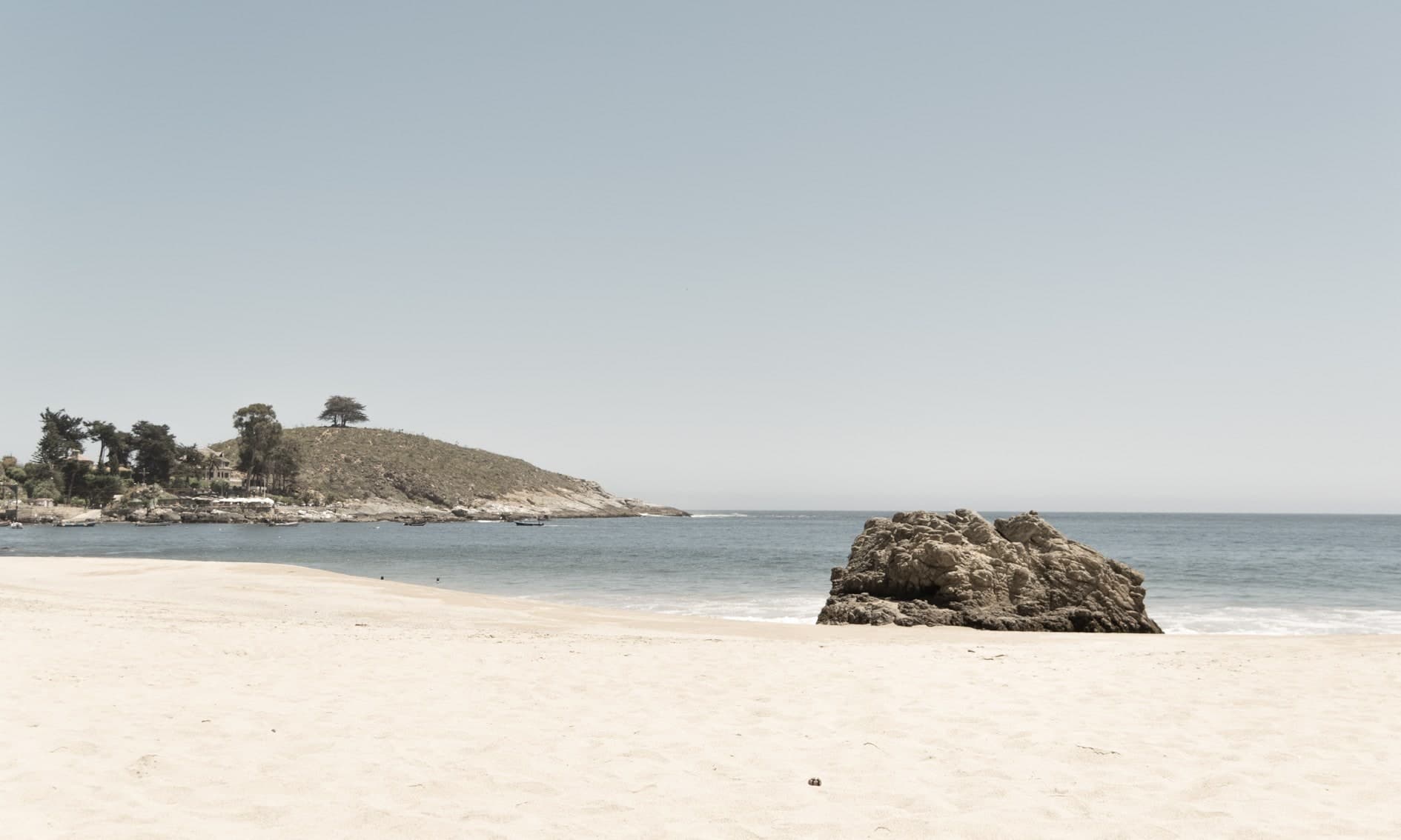 Playa de arena clara con gran roca al centro, península con árboles a la izquierda, cielo despejado. Agua #P20, fotografía de Cristian Letelier, edición limitada