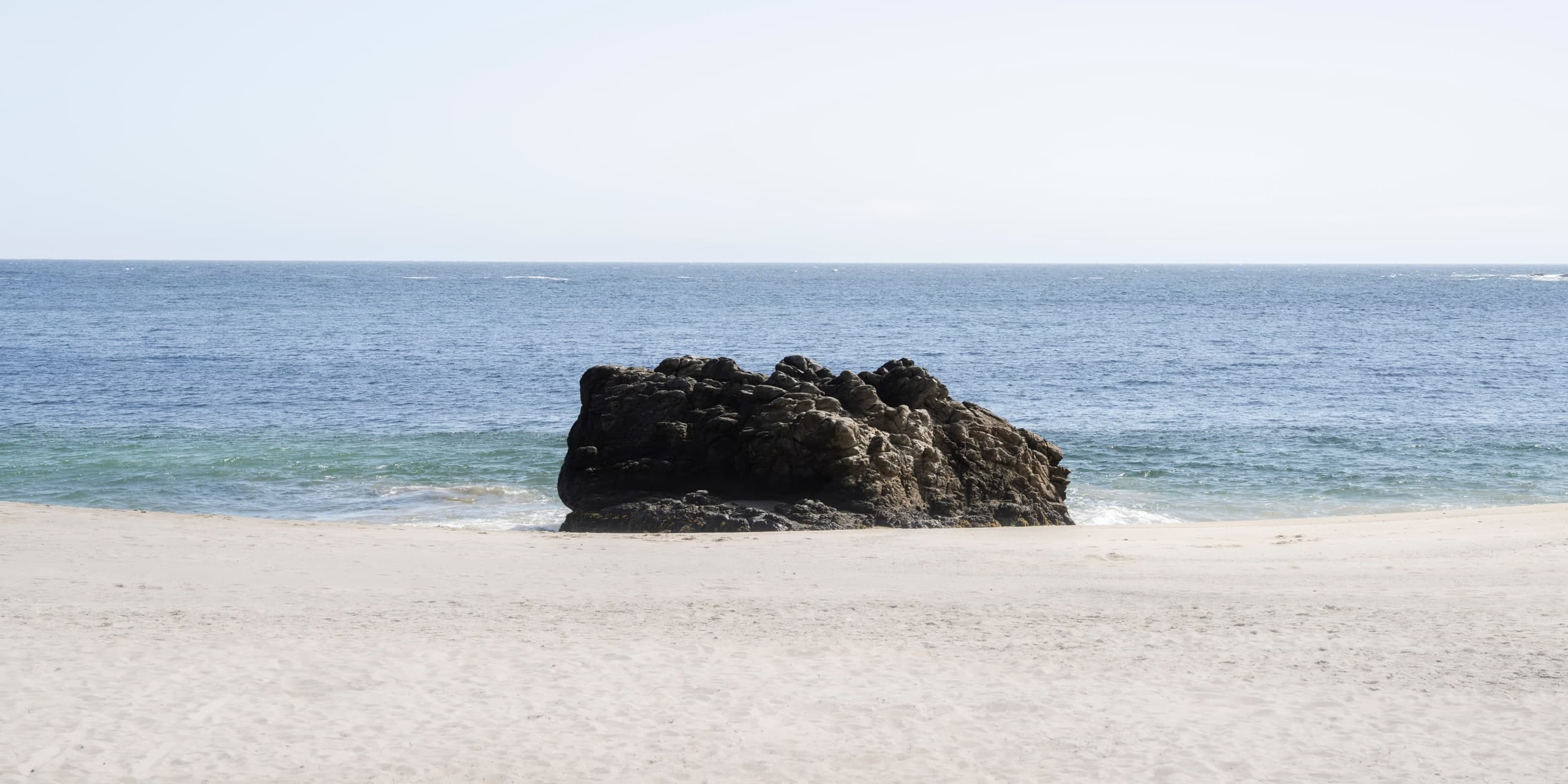 Playa de arena clara con una gran roca oscura en la orilla, mar azul en calma y cielo despejado; panorámica. Agua #P39, fotografía de Cristian Letelier, edición limitada