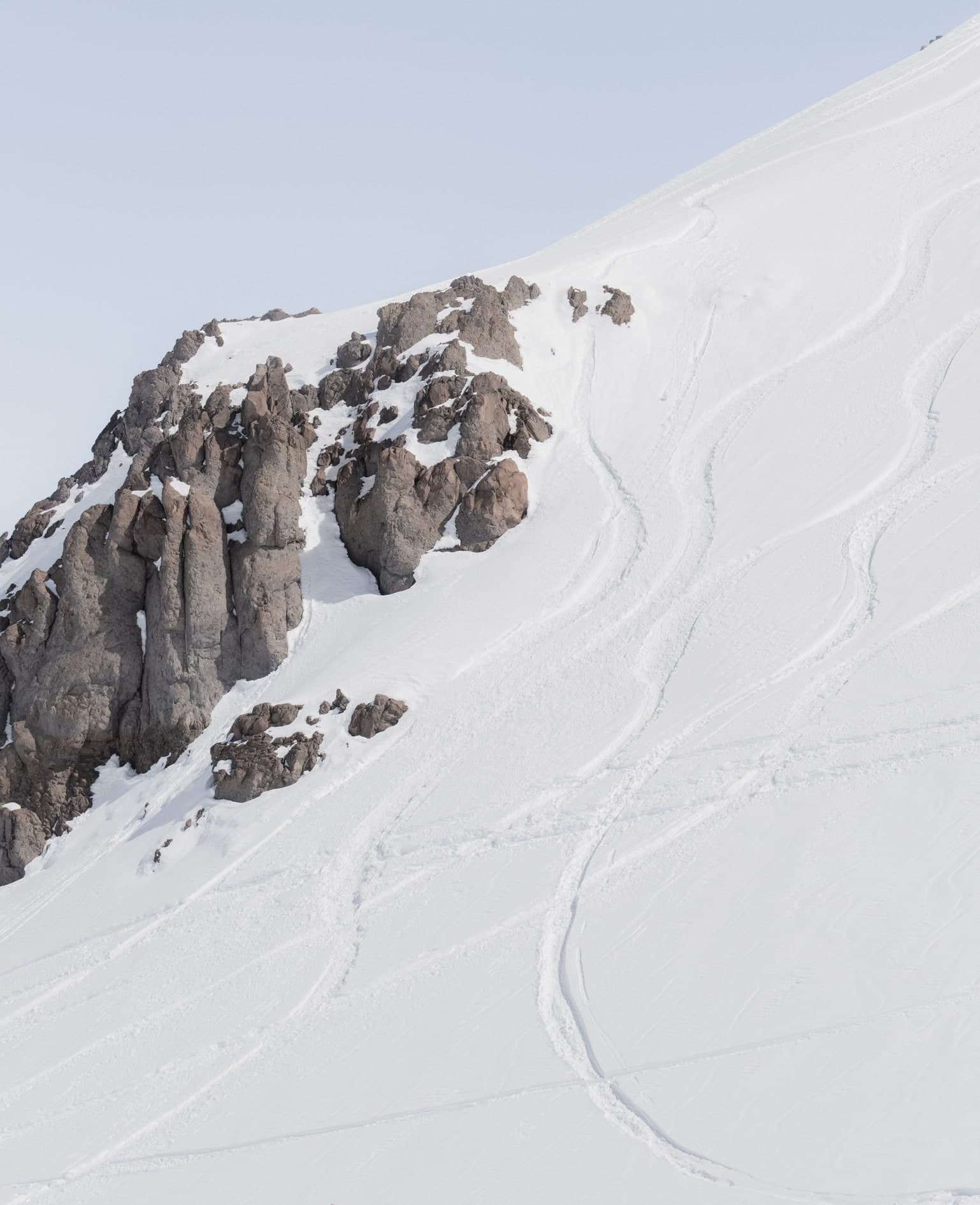 Vertical: ladera nevada con rocas oscuras a la izquierda y huellas de esquí curvas, cielo celeste. Aire #C7, fotografía de Cristian Letelier, edición limitada