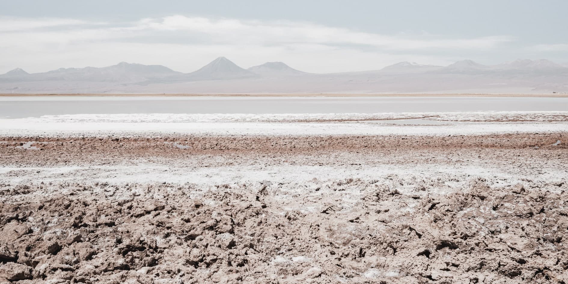 Salar con costra de sal marrón y blanca en primer plano, franja blanca y volcanes al fondo, cielo pálido. Fuego #D1, fotografía de Cristian Letelier, edición limitada