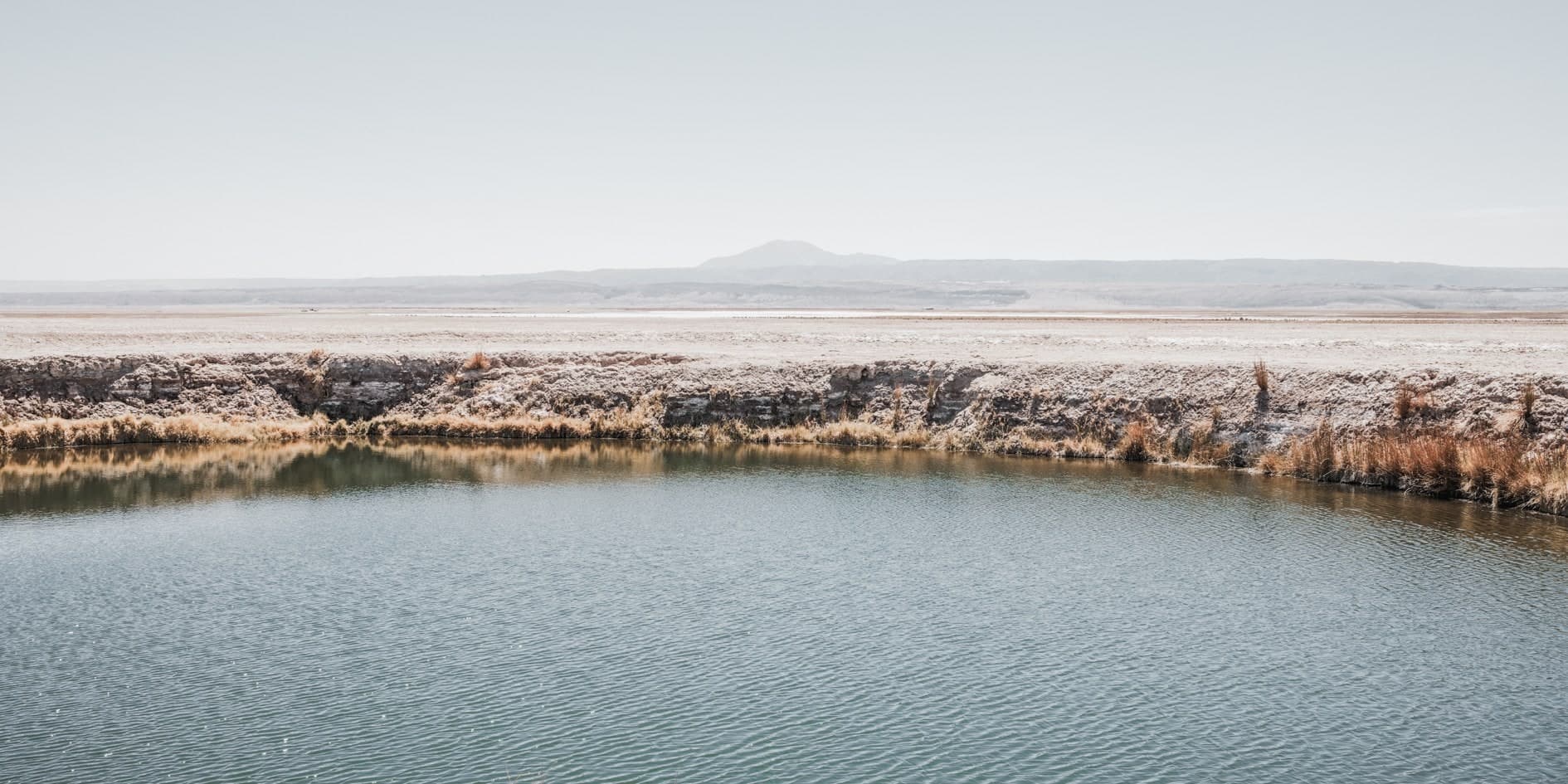 Laguna turquesa rodeada de orilla salina con pastos secos, planicie y cerro lejano al fondo. Fuego #D3, fotografía de Cristian Letelier, edición limitada