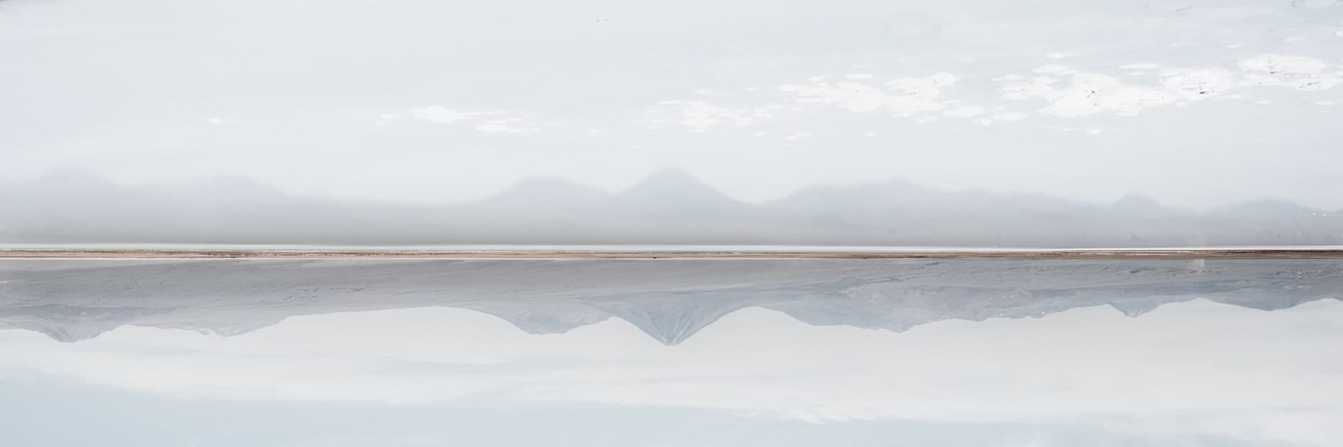 Panorámica de cerros y volcanes reflejados en agua quieta de salar, tonos gris-azul muy pálidos, casi abstracta. Fuego #D5, fotografía de Cristian Letelier, edición limitada