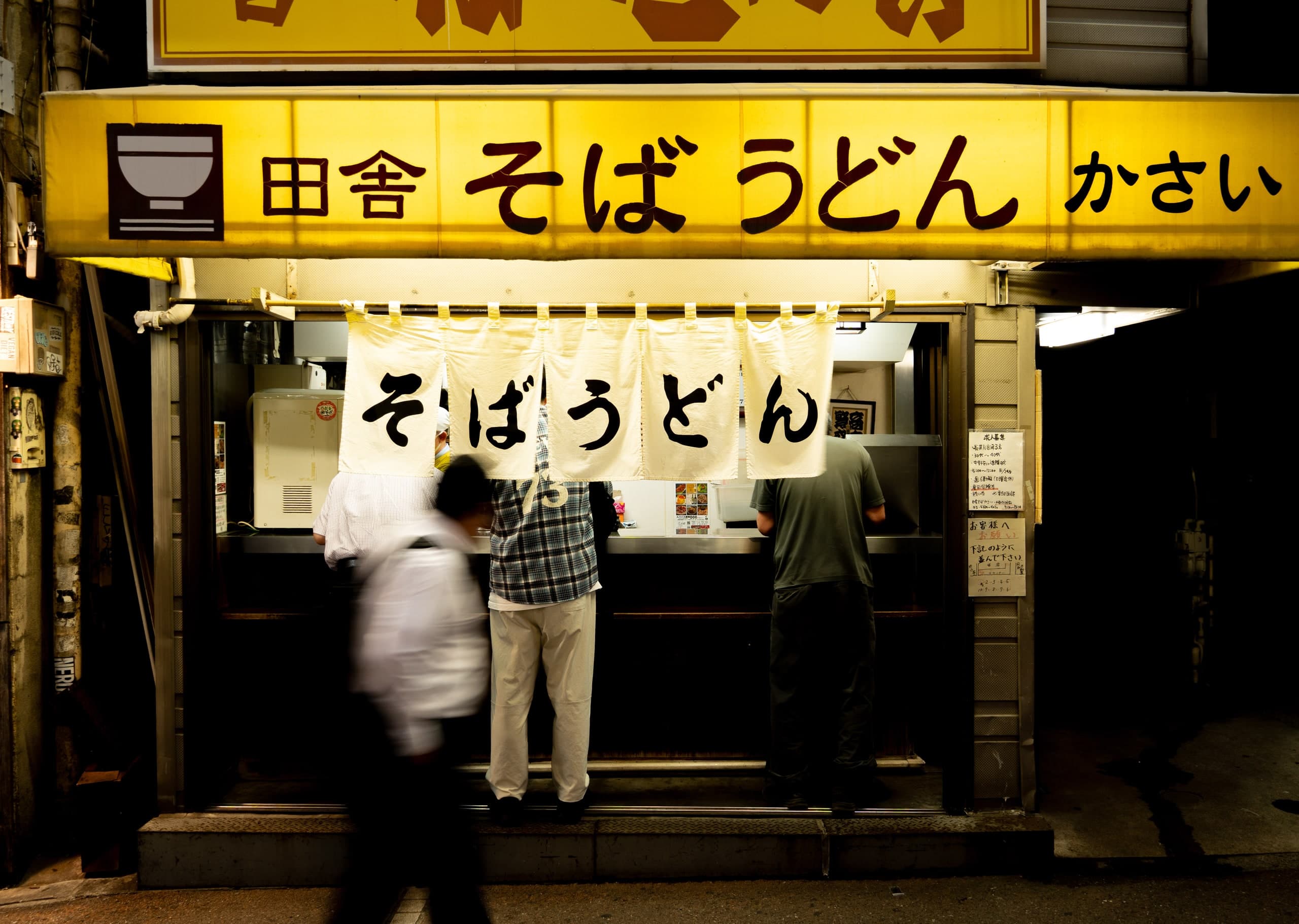 Puesto de soba y udon de noche: letrero amarillo, cortinas noren blancas y clientes en la barra, un peatón pasa movido. Japón #J1, fotografía de Cristian Letelier, edición limitada