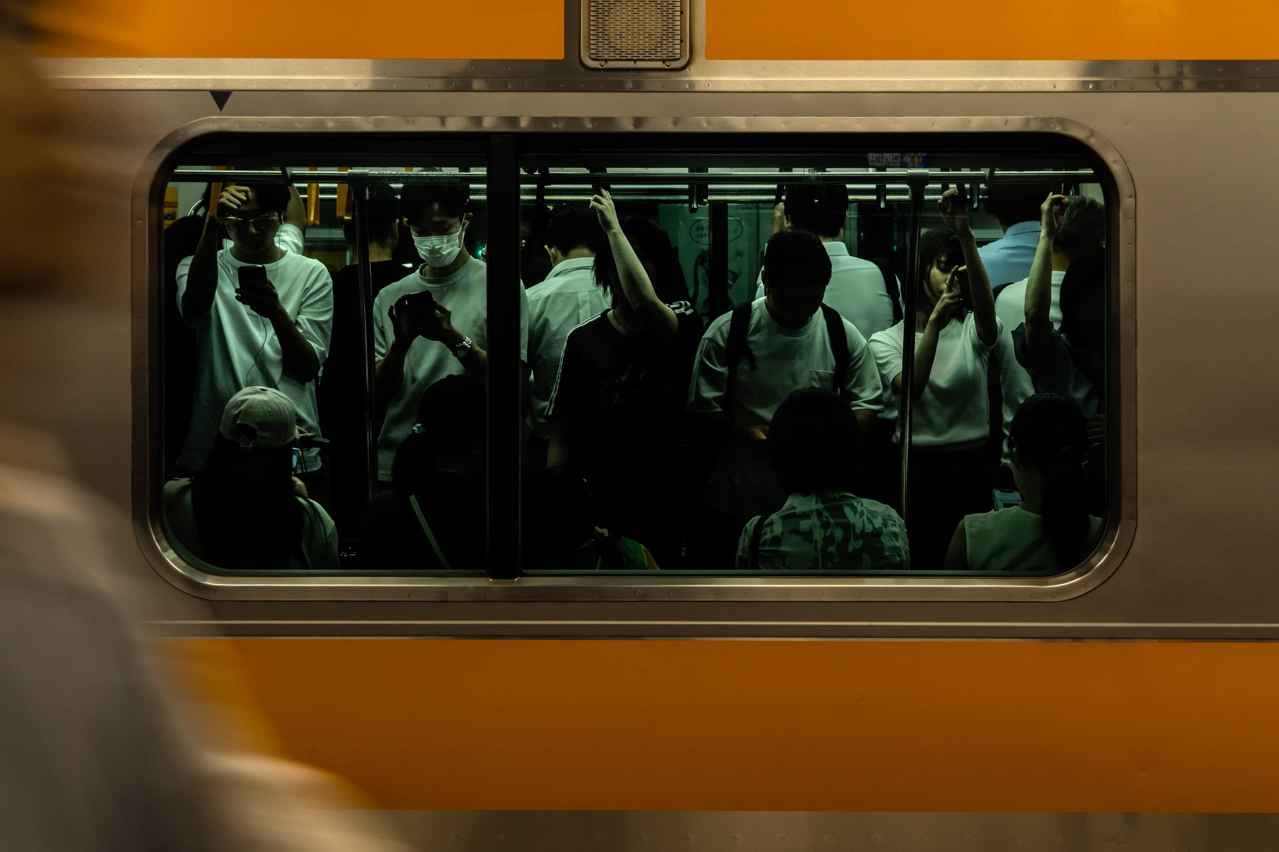 Ventana de un tren naranja repleto: pasajeros de pie mirando sus teléfonos, un andén borroso en primer plano. Japón #J2, fotografía de Cristian Letelier, edición limitada