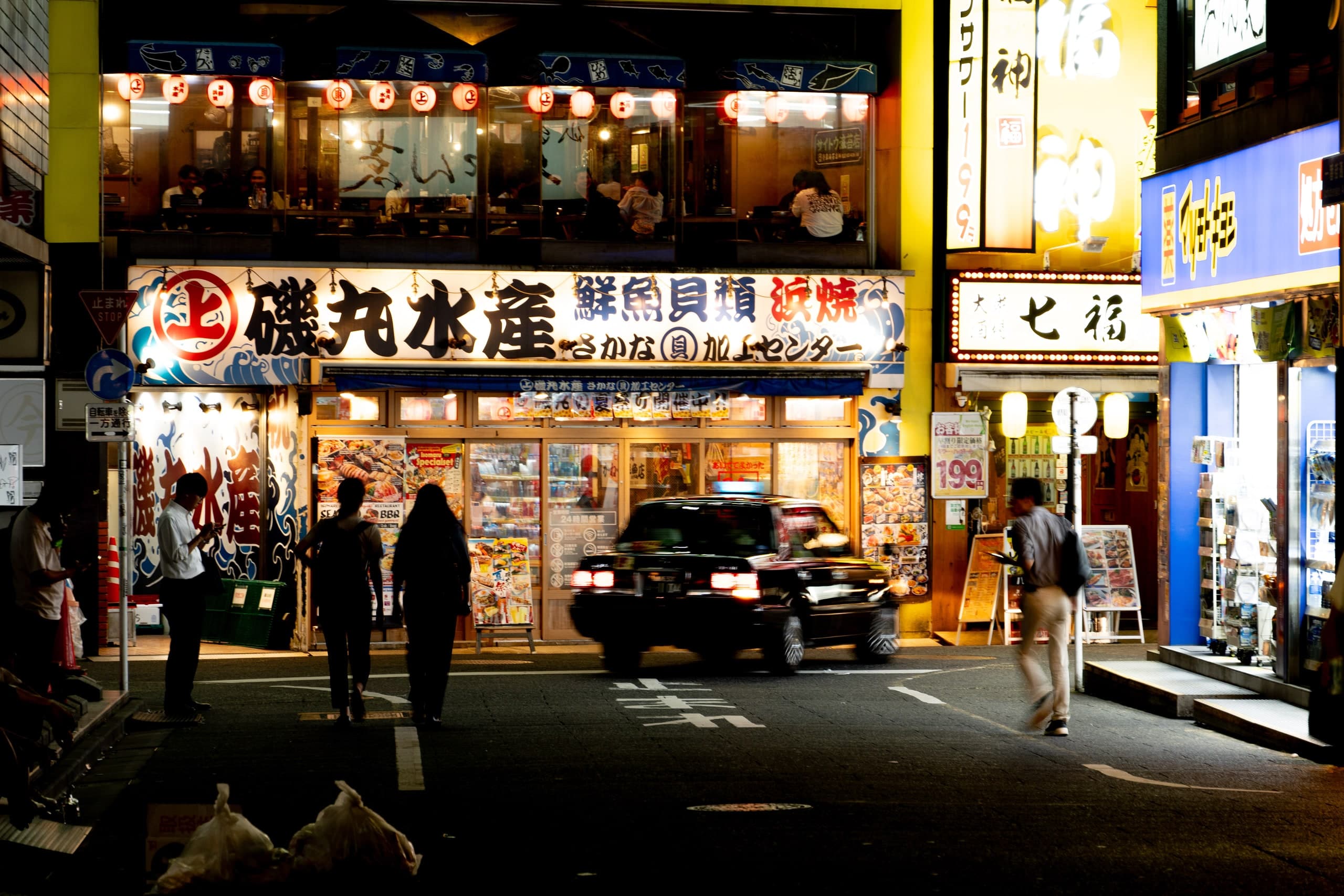 Calle de Tokio de noche: fachada de una pescadería iluminada con faroles rojos, un taxi negro y peatones. Japón #J3, fotografía de Cristian Letelier, edición limitada