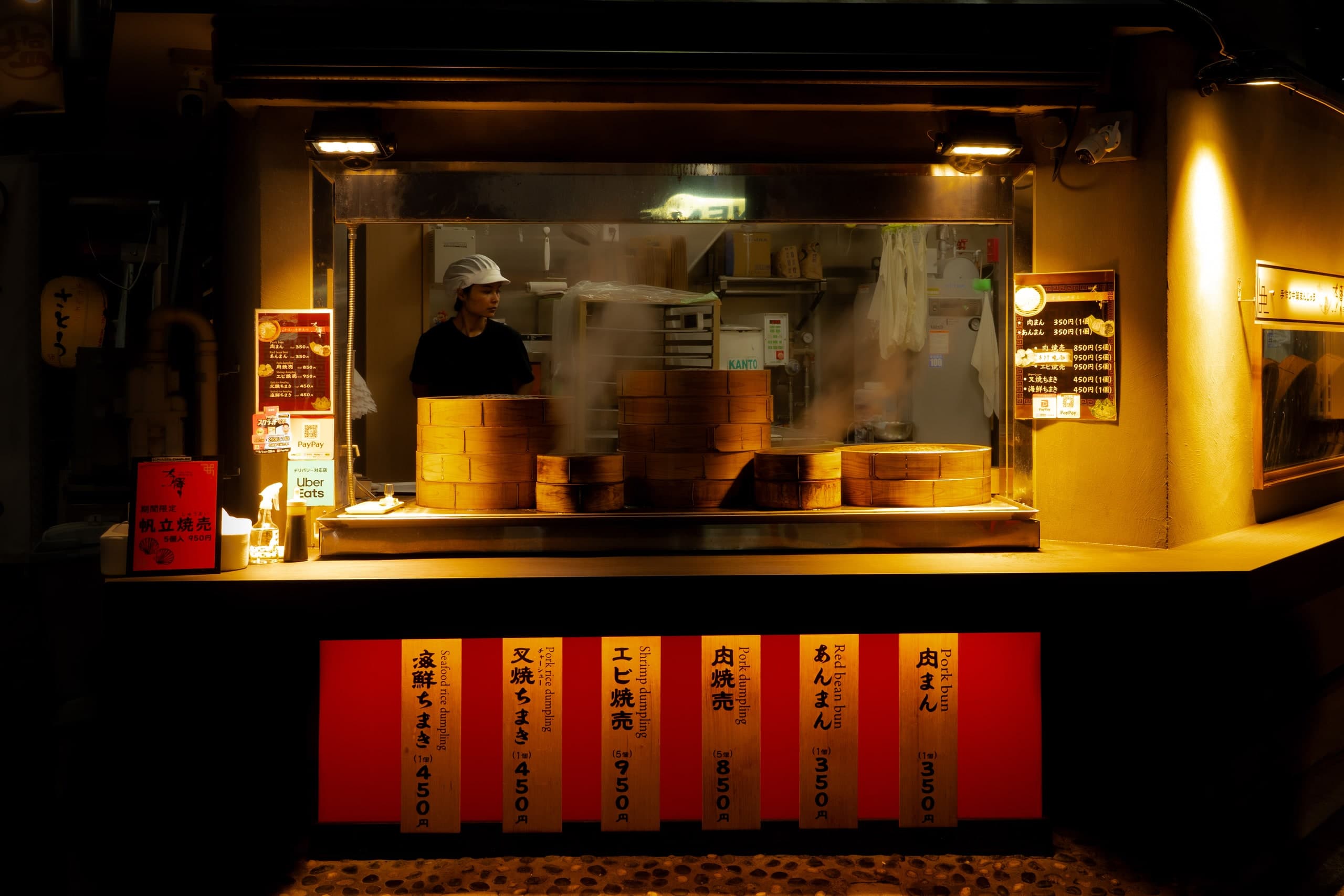 Puesto de dumplings de noche: cestas de bambú humeantes, una cocinera detrás y carteles de madera sobre rojo. Japón #J4, fotografía de Cristian Letelier, edición limitada