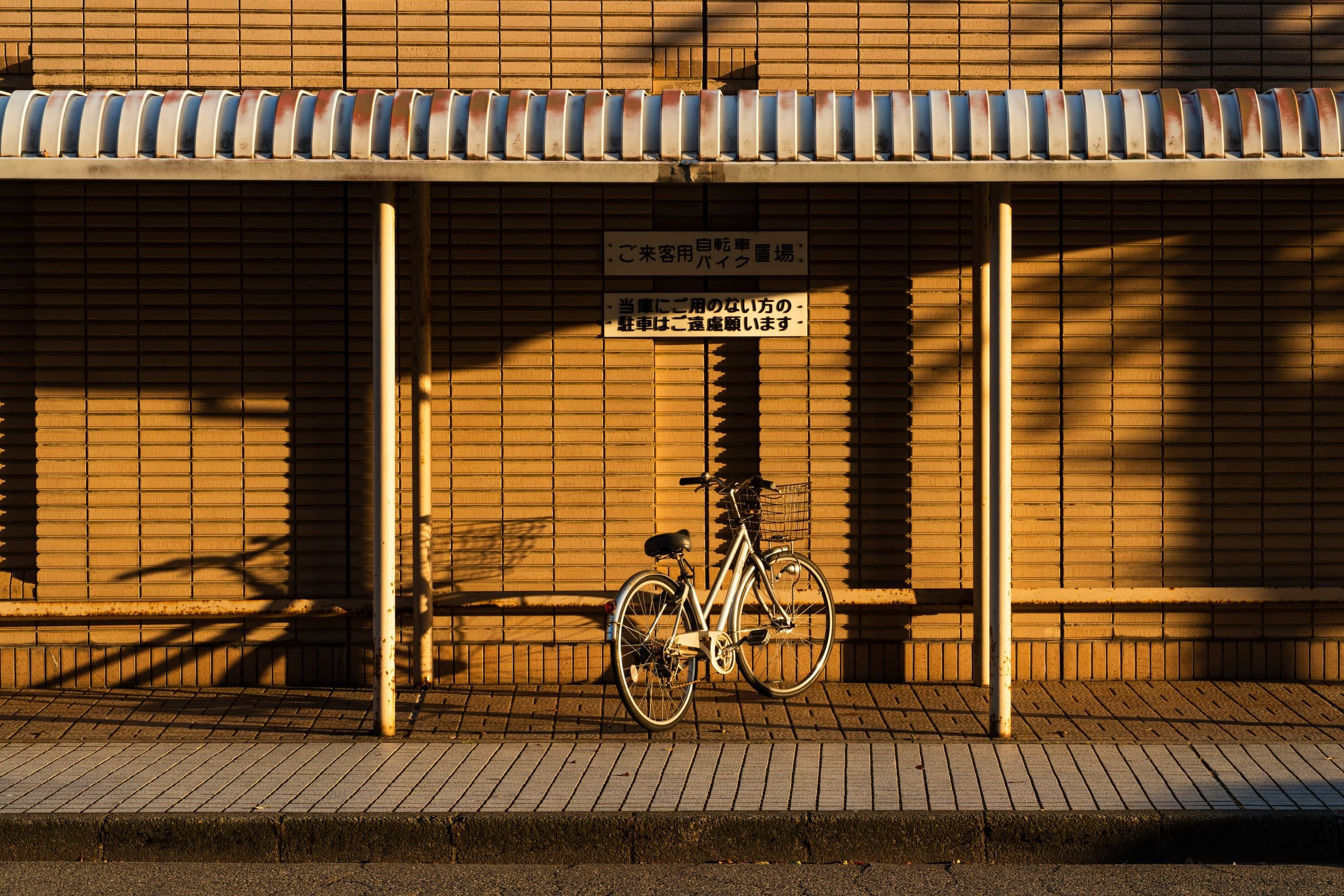 Bicicleta sola bajo un alero, contra un muro de ladrillo ocre con sombras largas del atardecer. Japón #J5, fotografía de Cristian Letelier, edición limitada