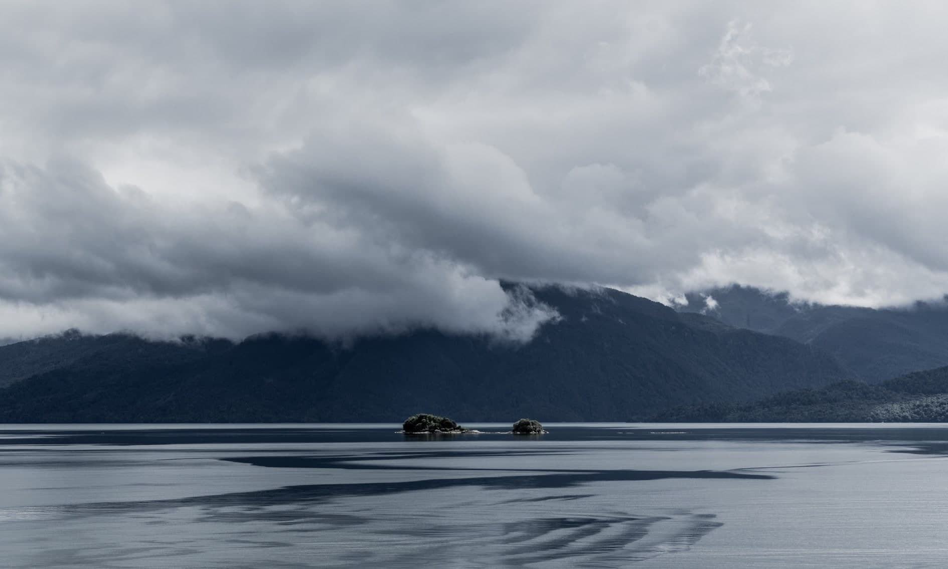 Lago calmo con dos rocas pequeñas, montañas y nubes bajas; color azul-gris. Tierra #S13, fotografía de Cristian Letelier, edición limitada