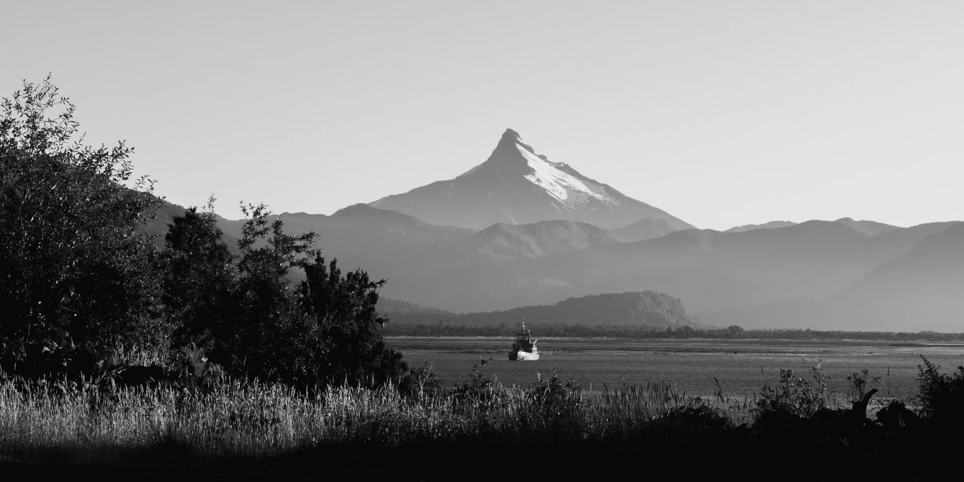 Volcán de cono nevado sobre lago con embarcación, arbustos en primer plano; blanco y negro. Tierra #S20, fotografía de Cristian Letelier, edición limitada